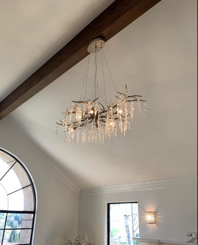 A branch-style crystal chandelier suspended from a vaulted ceiling beside an exposed beam, with a wall sconce lit below.