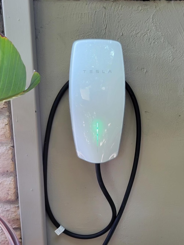 An EV wall connector mounted on an exterior wall with its charging cable coiled neatly beneath it.